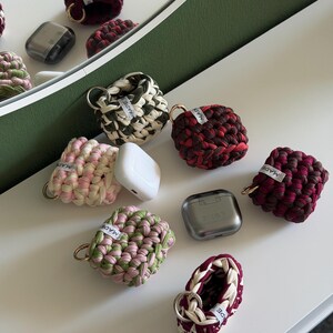 Puede incluir: Una colección de fundas cuadradas de tela tejida a ganchillo para auriculares inalámbricos, con varias combinaciones de colores que incluyen granate, verde oliva, rosa y crema. Cada funda incluye una pequeña anilla de metal dorado y una etiqueta blanca MADE.