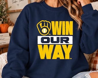 May include: A navy blue crewneck sweatshirt featuring a yellow and white graphic print. The design includes a baseball glove logo with a ball inside, followed by the bold text WIN OUR WAY in a distressed, weathered font style.