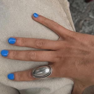 Könnte beinhalten: Ein silberner Ring mit einem großen, glatten, ovalen Mittelstück mit polierter Metalloberfläche. Der Ring wird am Ringfinger getragen und zeigt ein massives, abgerundetes Banddesign, das deutlich auf der Oberfläche des Fingers aufliegt.
