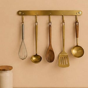 May include: A wall-mounted brass kitchen utensil rack featuring five hooks. Hanging from the hooks are a silver wire whisk, a brass ladle, a wooden spoon, a brass slotted spatula, and a brass ladle with a wooden handle accent.