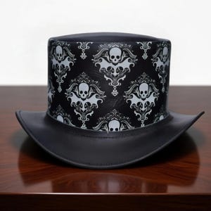 May include: A black top hat featuring a repeating white damask pattern of skulls and ornate scrollwork. The hat has a flat crown and a curved brim, with a smooth, solid black finish on the brim and a detailed graphic print on the cylindrical body.