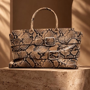 Könnte beinhalten: Eine strukturierte Handtasche mit einem braun-schwarzen Schlangenledermuster. Die Tasche verfügt über zwei horizontale Riemen auf der Vorderseite, die jeweils mit einer silberfarbenen Metallschnalle befestigt sind. Sie hat einen Tragegriff.