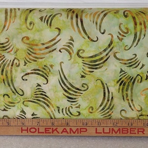 Op de afbeelding: Groen en bruin batik-stof met een draaiend patroon. De stof wordt gemeten met een houten liniaal waarop "Holekamp Lumber Co." staat.