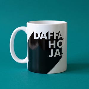 May include: A white ceramic mug featuring a large black geometric shape on the side. The text "DAFFA HO JA!" is printed in bold white capital letters across the black area. The mug has a curved handle on the left side and a smooth, glossy finish.