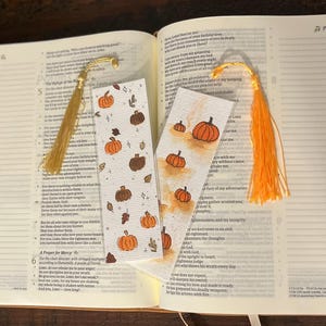 May include: Two rectangular paper bookmarks resting on an open book. One bookmark features a repeating pattern of orange and brown pumpkins with small stars. The other has a watercolor-style orange wash with pumpkin illustrations and a bright orange tassel.
