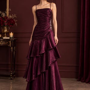 Könnte beinhalten: Ein bodenlanges, tiefburgunderrotes Kleid mit quadratischem Ausschnitt, dünnen Spaghettiträgern und einem eng anliegenden Oberteil mit horizontalen Raffungen. Der Rock ist mit kaskadierenden, asymmetrischen Rüschen und Perlen verziert.