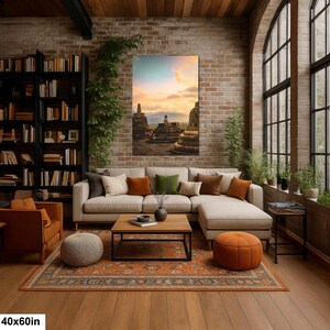 Op de afbeelding: Een rechthoekige wandkunstprint van 40 bij 60 cm met een zonsondergang van stenen stupa's en een Boeddhabeeld. Het kunstwerk hangt aan een bakstenen muur boven een beige hoekbank met oranje en groene sierkussens in een woonkamer.