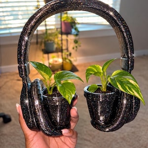 May include: A black glittery headphone-shaped planter set featuring two integrated pots, each containing a small green and yellow variegated pothos plant. The structure is connected by a curved headband and has a sparkling finish.