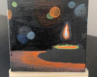 May include: A small square canvas painting on a wooden easel featuring a dark black background with a central orange flame, a glowing orange circular shape, and various small blue, green, and orange dots scattered across the composition.