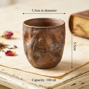 May include: A brown, textured ceramic cup with an irregular, organic surface. The cup has a diameter of 5.3 cm, a height of 5.5 cm, and a capacity of 100 ml. The exterior features a mottled, earthy finish with dark and light brown tones.