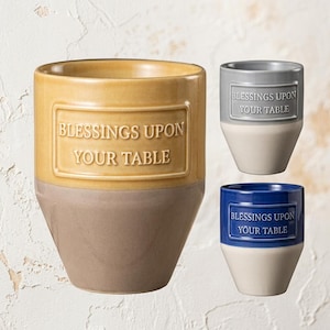 May include: Three ceramic cups with a two-tone design. The largest cup is yellow and tan, the top right is grey and white, and the bottom right is blue and white. Each cup features a rectangular plaque with the text BLESSINGS UPON YOUR TABLE.
