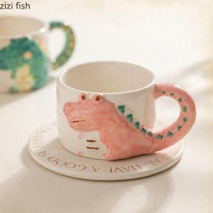 Pode incluir: Uma caneca de cerâmica branca com um design de dinossauro rosa em relevo que forma a alça, apoiada num pires branco a condizer com o texto "HAVING A GOOD DAY" gravado ao longo da borda. Uma segunda caneca desfocada é visível ao fundo.