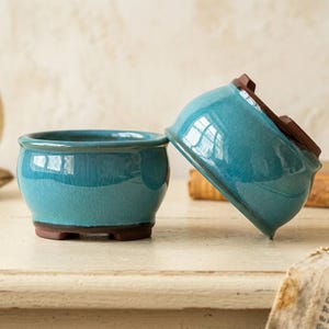 May include: Two round ceramic bonsai pots with a glossy turquoise glaze and exposed dark brown clay bases. One pot sits upright while the other is tilted on its side, revealing the unglazed brown underside and small feet on the bottom.