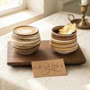 May include: Two rounded ceramic cups with horizontal brown and white swirl patterns. One cup contains a latte with heart-shaped foam art, while the other holds a dried pineapple slice. A rectangular brown card with the text a gift for you is included.