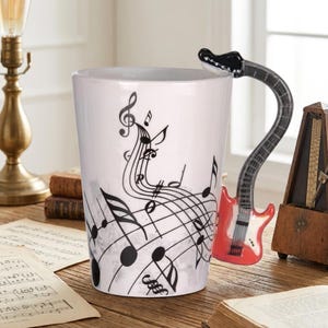 May include: A white ceramic mug featuring black musical notes and staff lines printed on the exterior. The handle is shaped like a red and black electric guitar, extending from the side of the mug to the top rim, providing a unique grip.