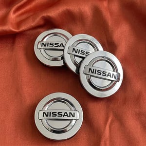 May include: Four circular silver-toned Nissan wheel center caps featuring a polished metallic finish. Each cap displays the Nissan brand name in black capital letters across a horizontal bar that bisects the circular emblem design.