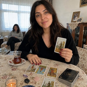 Op de afbeelding: Een persoon met lang donker haar in een zwarte trui met v-hals houdt een tarotkaart vast met een figuur die drie staven vasthoudt. Op de tafel liggen verschillende tarotkaarten, een kaartspel, een brandende kaars en kleine kristallen.