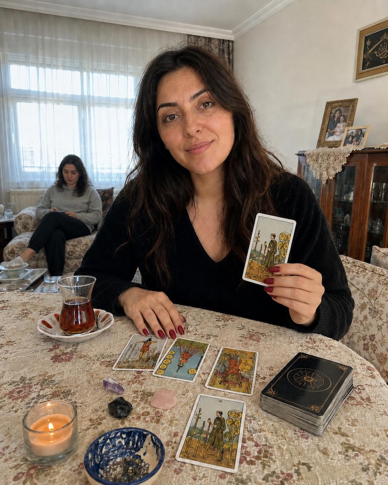 Op de afbeelding: Een persoon met lang donker haar in een zwarte trui met v-hals houdt een tarotkaart vast met een figuur die drie staven vasthoudt. Op de tafel liggen verschillende tarotkaarten, een kaartspel, een brandende kaars en kleine kristallen.