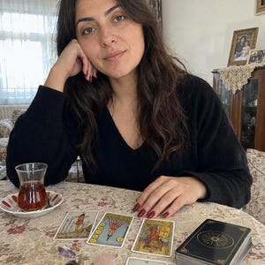 Op de afbeelding: Een zwarte V-hals trui gedragen door een persoon die aan een tafel zit met een tarotkaartspel, vier uitgestalde tarotkaarten, een glas donkere thee op een schoteltje, een klein brandend kaarsje in een glazen houder en drie kleine gepolijste kristallen.