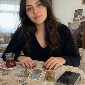 Op de afbeelding: Een persoon met lang donker haar in een zwarte trui met v-hals zit aan een tafel met vier uitgestalde tarotkaarten, een stapel kaarten, een klein theeglas, een brandende kaars in een houder, een blauw-wit kommetje en drie kleine kristallen.