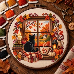 May include: A circular embroidery hoop containing a cross-stitch pattern of a black cat sitting on a plaid blanket by a window. The scene features autumn leaves, a lit candle, a steaming mug, a lantern, and pumpkins, with embroidery floss spools nearby.