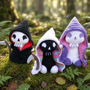 Puede incluir: Tres figuras de peluche tejidas a ganchillo con capuchas. La figura de la izquierda es negra con el interior de la capucha rojo y una guadaña. La central es negra con capucha blanca e interior arcoíris. La de la derecha es blanca con capucha morada y rosa.