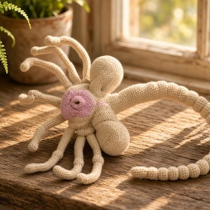 May include: A beige crocheted creature with eight long, slender legs, a rounded central body, and a long, segmented tail. The centre of the creature features a circular, light pink textured detail. The item is crafted from yarn.