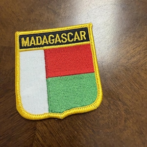 May include: A rectangular embroidered patch featuring the flag of Madagascar with a white vertical stripe on the left and red and green horizontal stripes on the right. The word MADAGASCAR is stitched in yellow on a black background at the top.