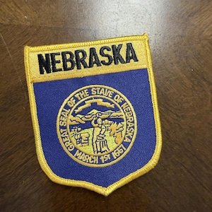 May include: A shield-shaped embroidered patch featuring the word NEBRASKA in black text on a gold background. Below, a blue field contains the Great Seal of the State of Nebraska, depicting a blacksmith, mountains, and the text MARCH 1ST 1867.