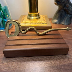 May include: A brass bottle opener shaped like a slithering snake with a textured, scale-like surface. The head of the snake forms the handle, while the looped tail section serves as the functional opening mechanism for removing bottle caps.
