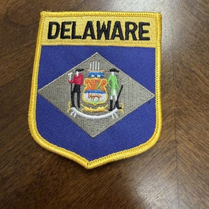 May include: A shield-shaped embroidered patch featuring a blue background with a yellow border. The word DELAWARE is stitched in black at the top. A gray diamond in the center displays a colorful state seal with two figures and a shield.