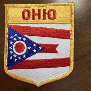 Puede incluir: Un parche bordado rectangular con un borde amarillo que presenta la palabra OHIO en letras rojas en la parte superior. Debajo, el diseño de la bandera del estado de Ohio muestra un triángulo azul con estrellas blancas y un círculo rojo, junto a franjas rojas y blancas.