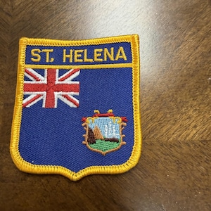 May include: A shield-shaped embroidered patch with a blue background and a yellow border. The text ST. HELENA is stitched in yellow at the top. It features a Union Jack in the upper left corner and a colorful coat of arms in the lower right.