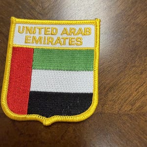 May include: A shield-shaped embroidered patch featuring the flag of the United Arab Emirates with a vertical red stripe on the left and horizontal green, white, and black stripes on the right. The text UNITED ARAB EMIRATES is stitched in gold.