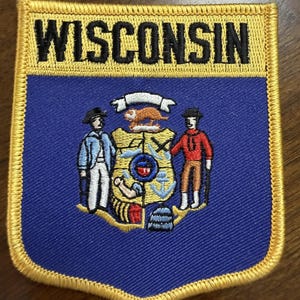May include: A rectangular embroidered patch with a yellow border and the word WISCONSIN in black text at the top. The lower section features a blue background with the state seal, depicting two figures flanking a shield with a badger and tools.
