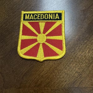 May include: A shield-shaped embroidered patch featuring the flag of North Macedonia. The design includes a central yellow sun with eight radiating rays on a red background, topped by a black horizontal bar with the word MACEDONIA in yellow.