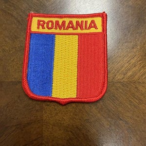 May include: A shield-shaped embroidered patch featuring the vertical tricolor flag of Romania in blue, yellow, and red. The top section contains a yellow horizontal bar with the word ROMANIA stitched in red capital letters, all bordered by red.