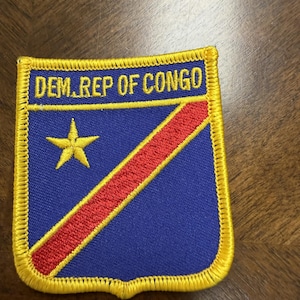 May include: A rectangular embroidered patch featuring the flag of the Democratic Republic of the Congo with a blue background, a diagonal red stripe, and a yellow star. The text DEM. REP OF CONGO is stitched in yellow at the top with a yellow border.
