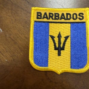 May include: A rectangular embroidered patch featuring the flag of Barbados with a yellow border. The design includes two blue vertical stripes flanking a central yellow stripe with a black trident symbol. The word BARBADOS is in black text.