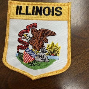 Op de afbeelding: Een schildvormige geborduurde patch met het woord ILLINOIS in zwarte letters op een gele achtergrond. Het midden toont een Amerikaanse zeearend op een rots met een rood lint met NATIONAL UNION STATE SOVEREIGNTY en een opkomende zon.