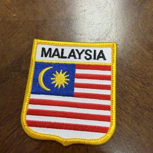 May include: A rectangular embroidered patch featuring the flag of Malaysia with a yellow border. The design includes a blue canton with a yellow crescent and a 14-pointed star, alongside red and white horizontal stripes. The word MALAYSIA is stitched in black capital letters on a white background at the top.