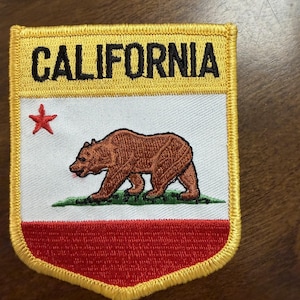 May include: A shield-shaped embroidered patch featuring the California state flag design. It displays the word CALIFORNIA in black text on a yellow background, a white field with a brown bear, a small red star, and a solid red horizontal stripe.