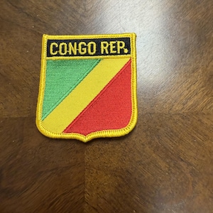 May include: A shield-shaped embroidered patch featuring the flag of the Republic of the Congo with diagonal green, yellow, and red stripes. The top section has a black horizontal bar with the yellow text CONGO REP. and a yellow border.