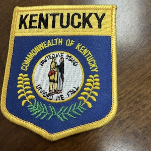 May include: A shield-shaped embroidered patch featuring the Kentucky state seal on a blue background with a yellow border. The text KENTUCKY appears at the top, with COMMONWEALTH OF KENTUCKY, UNITED WE STAND, and DIVIDED WE FALL embroidered in black.
