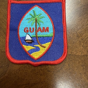 May include: A shield-shaped embroidered patch featuring a blue background with a red border. The center displays an oval emblem with a palm tree, a small sailboat on blue water, a yellow sandy shore, and the red text GUAM printed across the middle.