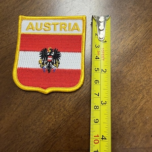 May include: A shield-shaped embroidered patch featuring the Austrian flag with a black eagle emblem and the word AUSTRIA in yellow text. A yellow measuring tape placed next to the patch shows it measures approximately 3 inches in height.