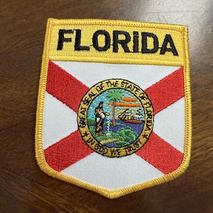 May include: A rectangular embroidered patch featuring the word FLORIDA in black text on a yellow background. Below, a white field with a red saltire cross displays the circular Great Seal of the State of Florida, depicting a person, sun, and water.