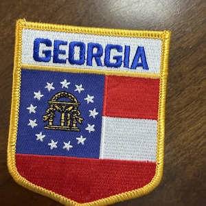 May include: A shield-shaped embroidered patch featuring the word GEORGIA in blue text on a white background. Below, a blue field contains a gold state seal surrounded by white stars, next to horizontal red and white stripes, all with a gold border.