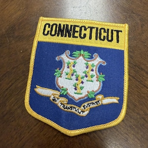 May include: A shield-shaped embroidered patch featuring the state of Connecticut. The top yellow bar displays the word CONNECTICUT in black. The blue field contains a white shield with three grapevines and a white ribbon with the text QUI TRANSTULIT SUSTINET.