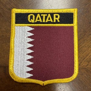 May include: A shield-shaped embroidered patch featuring the flag of Qatar with a maroon field and a white serrated vertical band on the left. The top section is a black horizontal bar with the word QATAR in yellow text, all bordered in yellow.